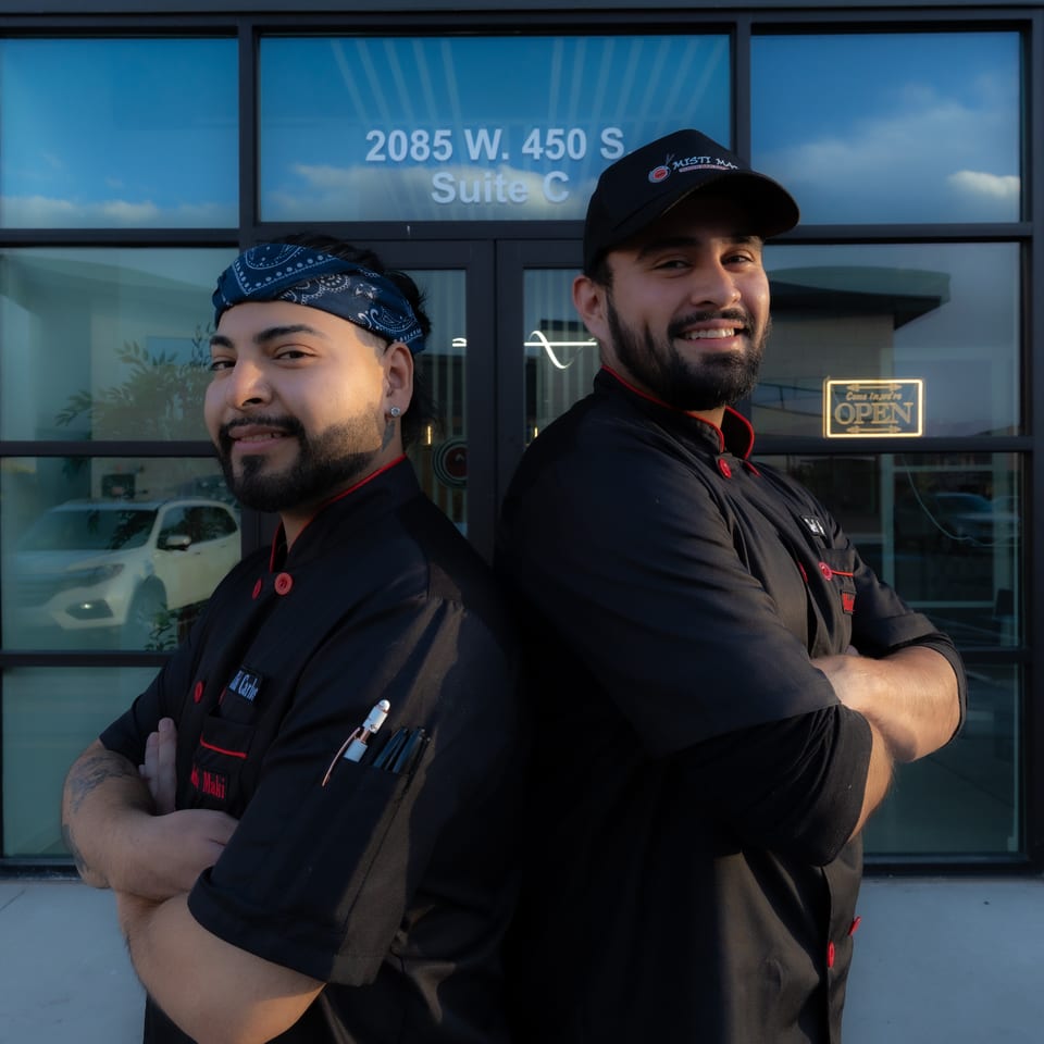 Two smiling chefs in black uniforms stand back-to-back outside a restaurant with an "OPEN" sign.