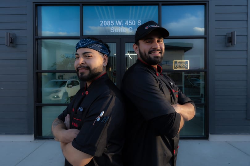 Two smiling chefs in black uniforms stand back-to-back outside a restaurant with an "OPEN" sign.