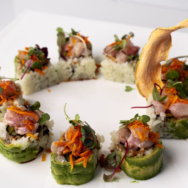 Eight avocado-wrapped sushi rolls with fish, microgreens, and shredded carrots on a white plate.