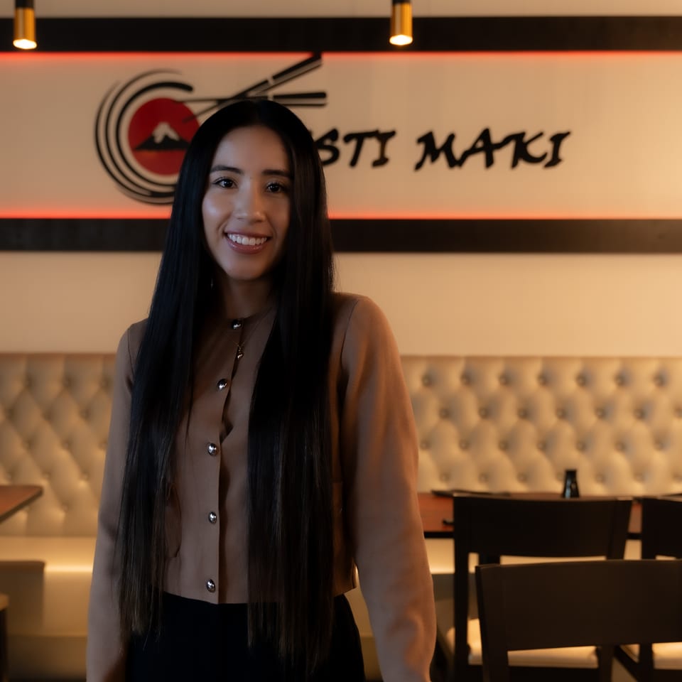 Smiling woman with long dark hair standing in a restaurant with a "Sosti Maki" sign.