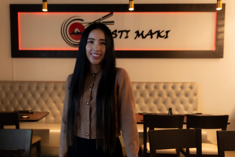 Smiling woman with long dark hair standing in a restaurant with a "Sosti Maki" sign.